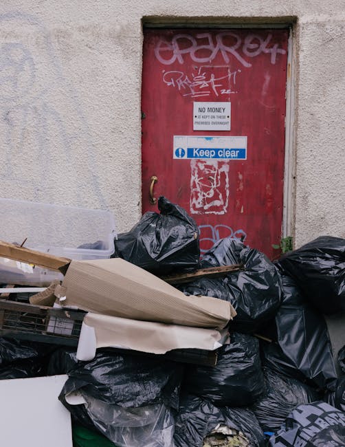 A large collection of black rubbish bags, some torn and partially open, are piled in front of a red metal door embedded in a beige concrete wall. The bags appear to contain various waste materials and