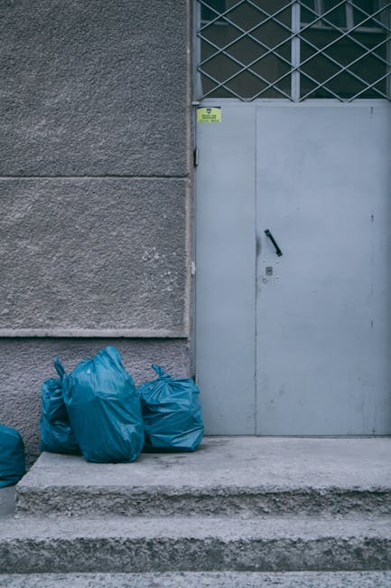 A group of five blue rubbish bags made of plastic are positioned on a concrete step in front of a grey metal door with a black handle and padlock, set within a wall that features a textured, rough gre